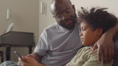 Afrikalı Amerikalı bir baba küçük oğlunu kucaklıyor ve evdeki kanepede birlikte otururken dijital tablette onunla bir şeyler tartışıyor.