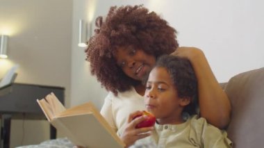 Küçük Afro-Amerikalı çocuk evde kanepede birlikte otururken, sevgi dolu annesiyle birlikte kitap okuyor ve elma yiyor.
