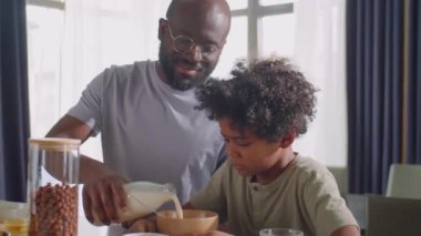 Neşeli Afrikalı Amerikalı baba küçük oğlu için mısır gevreğiyle kaseye süt dolduruyor, sonra portakal suyu içiyor ve kahvaltıda çocukla sohbet ediyor.