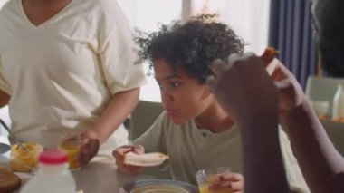 Afro-Amerikan baba ve küçük oğlu evde kahvaltı hazırlarken sandviç yiyor, meyve suyu içiyor ve annesiyle konuşuyor.