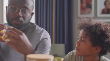 Küçük Afrikalı Amerikalı çocuk kahvaltıda kahvaltı ederken babasıyla konuşuyor ve kahvaltı yapıyor.