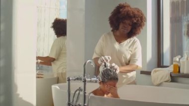 Afro-Amerikalı anne evdeki banyoda şampuanla küçük oğlunun saçını yıkıyor.