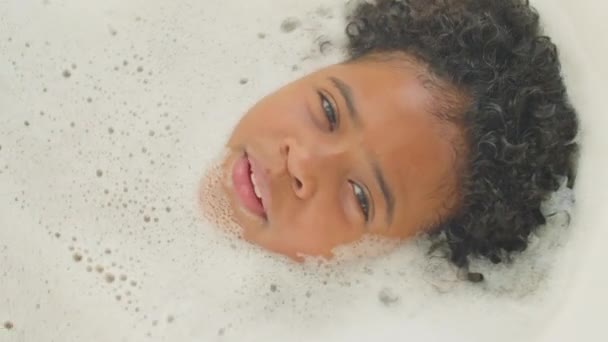 自上而下拍摄的非洲裔美国小男孩躺在泡着的洗澡水里 一边看着相机一边唱歌一边享受着洗澡时 — 图库视频影像