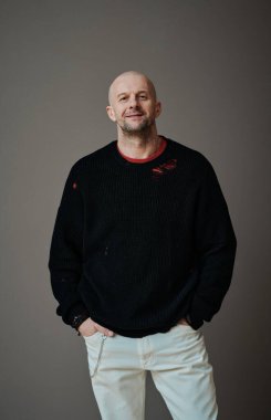 Dikey orta boy, kendine güvenen, olgun, beyaz bir adamın gri duvarın arkasında dikilip kameraya bakarken, moda konseptini yükseltirken.