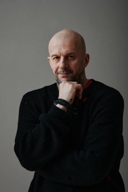 Siyah süveter giyen, gri arka planda kameraya bakan, kendine güvenen beyaz bir adamın dikey orta stüdyo portresi.