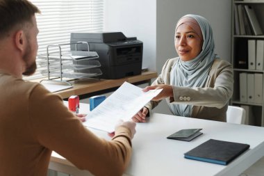 Elçilikte çalışan, tesettürlü, modern Müslüman bir kadın. Beyaz adamın önünde oturuyor. Vize onayı aldıktan sonra ona belgeler veriyor.