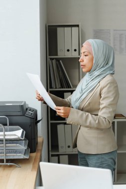Yazıcının önünde çalışan ve kağıt dökümanlarını kontrol eden modern, genç Müslüman kadının dikey yan görünüşü