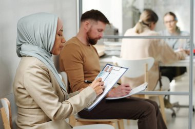Başörtülü genç beyaz erkek ve Müslüman kadın vize hizmetleri acentesinde sandalyelerde oturmuş randevu beklerken başvuru formlarını dolduruyorlar.