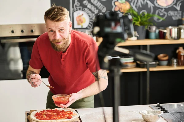 髭を生やした白人男性がピザのレシピビデオを撮影フードブログに自家製トマトソースを生地に塗り カメラで話す — ストック写真
