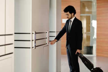 Siyah takım elbiseli genç işadamı ofis binasında duran asansörün düğmesine basıyor.