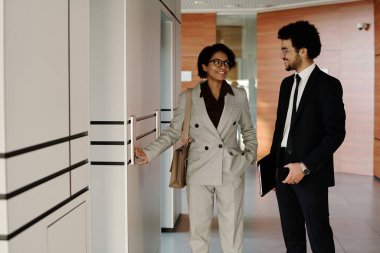 Genç iş arkadaşları ofis koridorunda asansör beklerken birbirleriyle konuşuyorlar.