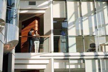 Genç iş adamları modern ofis binasının balkonunda dikilirken dijital tablet kullanıyorlar.