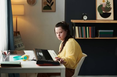 Down sendromlu genç beyaz bir kız üzerinde elektronik piyanoyla oturmuş tuşlara dokunarak bir şeyler düşünüyor.