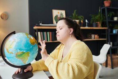 Down sendromlu düşünceli genç beyaz kadın evinde oturmuş dünya haritası okuyor seyahat noktalarını düşünüyor.