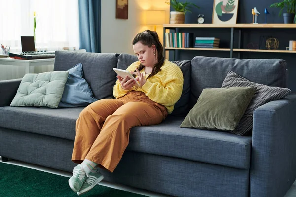 stock image Portrait of modern girl with Down syndrome sitting on sofa in living room at home using digital tablet