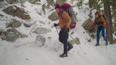 Bir çift turist, kış günü dağlarda birlikte yürüyüş yaparken yürüyüş direkleri ve sırt çantalarıyla dik patikada yürüyorlar.