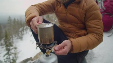 Erkek turist taşınabilir gaz ocağını açıyor ve kış kampı sırasında dağlarda sıcak içecek hazırlarken üzerine buz konmuş çelik bardak koyuyor.