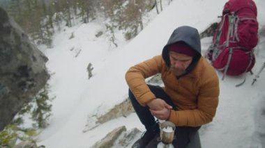 Erkek turist, kış yürüyüşü sırasında dağlarda kamp yaparken taşınabilir gaz ocağında çelik bardakta buzu eritiyor.