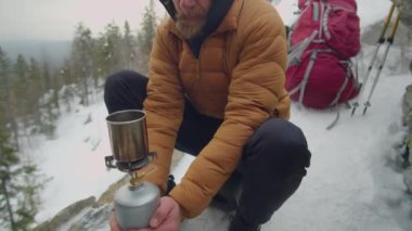 Adam ellerini gaz sobasıyla ısıtırken soğuk kış günlerinde dağlardaki kamp alanında çelik bardakta kaynar su içiyor.