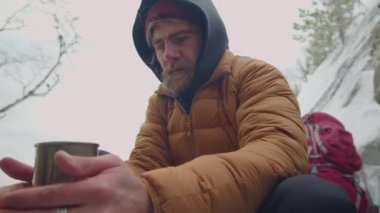 Erkek turist, dağlarda kamp yerinde otururken ve soğuk kış günlerinde ısınmaya çalışırken elinde sıcak çayla çelik bardak tutuyor.