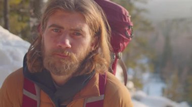 Sırt çantalı uzun saçlı erkek turistin portresi dağda duruyor ve kış günü kameraya poz veriyor.