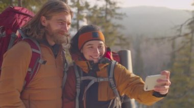 Sırt çantalı mutlu çift, dağın tepesinde durup akıllı telefonlarıyla selfie çekiyorlar ve kış günü doğa yürüyüşü yaparken bunu tartışıp duruyorlar.
