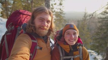 Sırt çantalı mutlu turist çiftin POV 'u, kış yürüyüşü sırasında dağın tepesinde selfie çekerken kameralara gülümsüyor ve el sallıyorlar.
