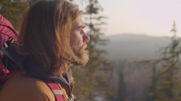 山の上に立ち 冬の森の景色を楽しむバックパック付きの長い毛髭の男のラックフォーカスショット — ストック動画