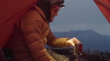 Erkek turist, kışın dağın tepesinde kamp çadırında otururken portatif gaz sobasıyla çay hazırlıyor ve ısınıyor.