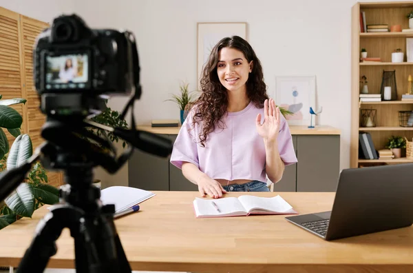 stock image Attractive young woman sitting at desk in front of camera on tripod starting video for blog with greeting her followers