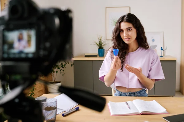 stock image Young woman with long curly hair sitting at desk in front of camera speaking about experience of using menstrual cup while recording video for blog