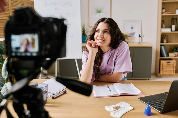 stock image Attractive young Caucasian woman with long curly hair sitting at desk in front of camera speaking about something while recording video for blog