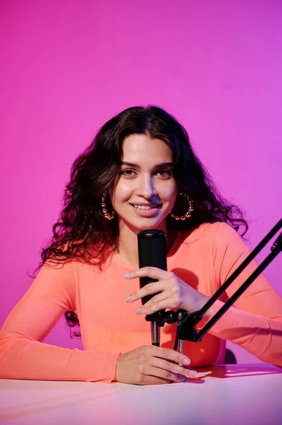 Stock image Vertical portrait of joyful female influencer sitting at desk in studio in pink neon light whispering in microphone for ASMR vlog