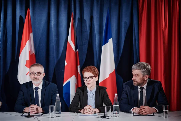 stock image Group of politicians sitting at table during press conference and answering media questions with flags in background