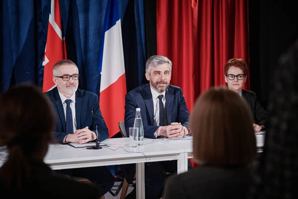 stock image Mature man with gray hair answering questions of journalists together with his colleagues while sitting at table at press conference