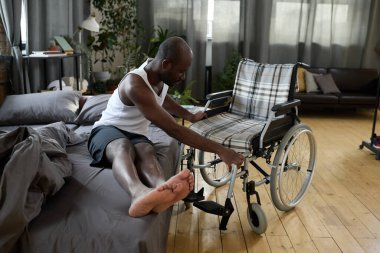 Özürlü bir Afrikalı, evinde tekerlekli sandalyesine yaslanarak yataktan kalkmaya çalışıyor.