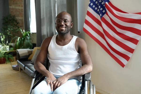 非洲裔美国人坐在轮椅上靠着房间墙上的美国国旗对着镜头微笑的画像 — 图库照片