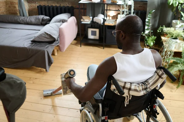 アフリカ系アメリカ人の男性が車椅子に座って掃除機でリビングの床を掃除している様子 — ストック写真