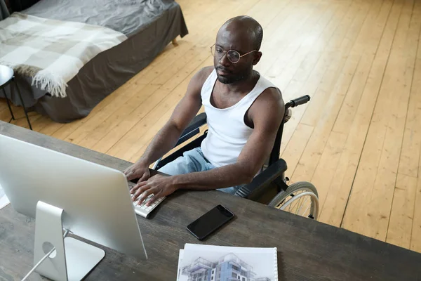 アフリカ系アメリカ人男性の車椅子に座ってコンピュータのモニターの前に座って 自宅でのオンライン作業中にコンピュータのキーボードを入力するの高い角度ビュー — ストック写真
