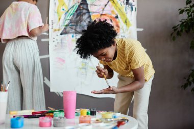 Afrikalı küçük çocuk elinde boya fırçasıyla boya karıştırırken kız kardeşiyle birlikte büyük bir resim çiziyor.