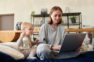 Genç bir kadın bebeğiyle yatakta oturuyor ve bilgisayarını internet iletişimi için kullanıyor.