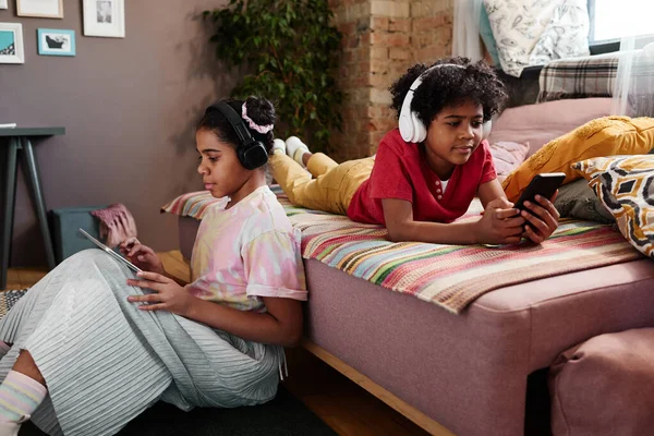 孩子们把业余时间花在家里买小玩艺儿上 他们坐在无线耳机里 用数字平板电脑和智能手机 — 图库照片