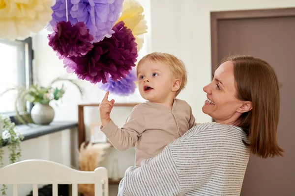 Young mother holding her baby on hands and smiling while he playing with paper decoration hanging above them