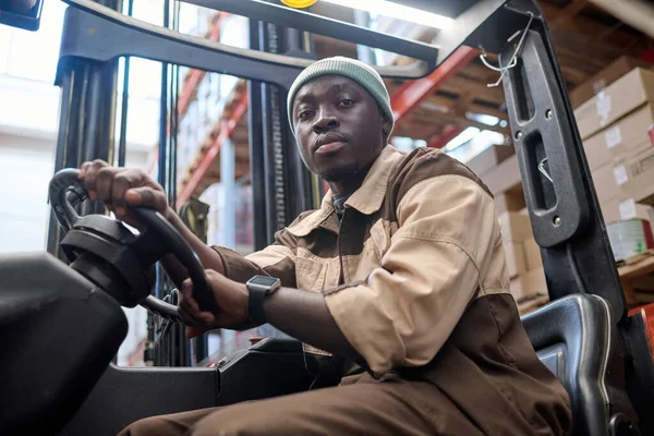 Portrait of African american driver looking at camera while driving forklift at warehouse