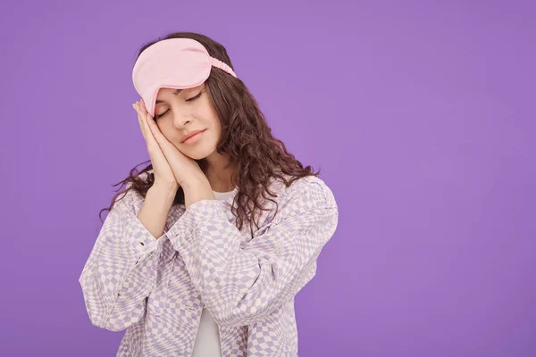 Portrait of young woman in sleep mask on her head with closed eyes pretending to sleep on purple background