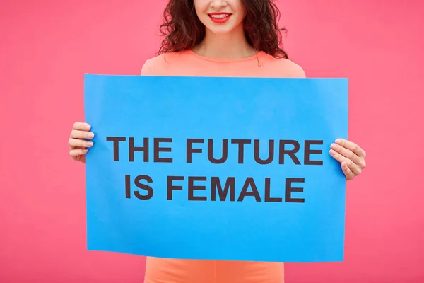 Close-up of young smiling woman holding The future is female poster to express her opinion in female rights isolated on pink background