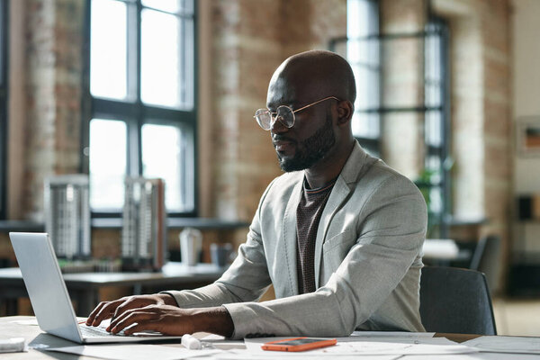 African serious businessman in eyeglasses sitting at his workplace at office and working on laptop