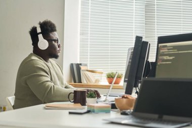 Kablosuz kulaklık takan Afrikalı yazılım geliştiricisi işyerinde oturuyor ve online iş için laptop kullanıyor