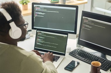 Kablosuz kulaklıklı Afrikalı bir adamın iş yerindeki bilgisayarına kod yazması.