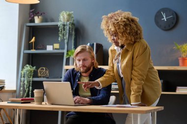 Kıvırcık saçlı Afrikalı iş kadını dizüstü bilgisayara bakıyor ve ofisteki meslektaşıyla online sunumu tartışıyor.